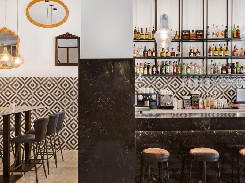 Modern bar interior with patterned tiles, dark marble counter, wooden stools, and hanging lights.