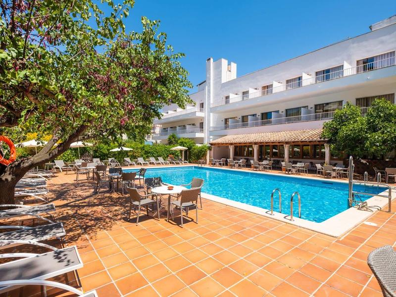 Hotelzwembad met blauw water, ligstoelen, bomen en een meerlagig gebouw onder een heldere lucht.