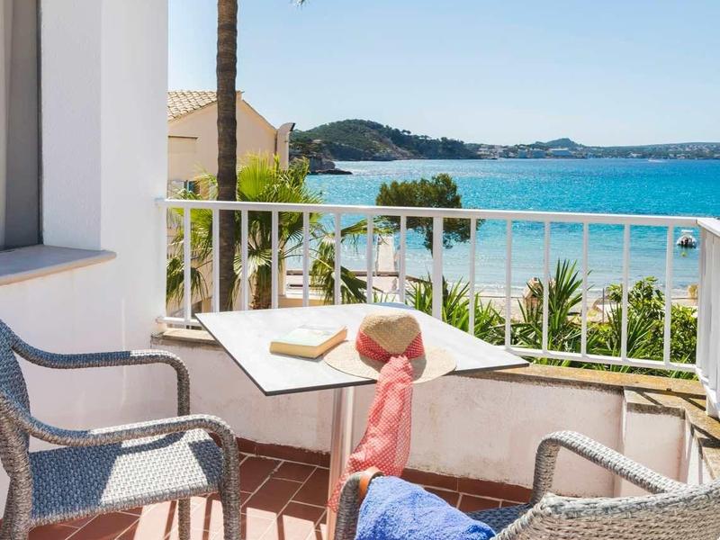 Balkon met stoelen, tafel en uitzicht op zee en palmbomen onder een blauwe lucht.