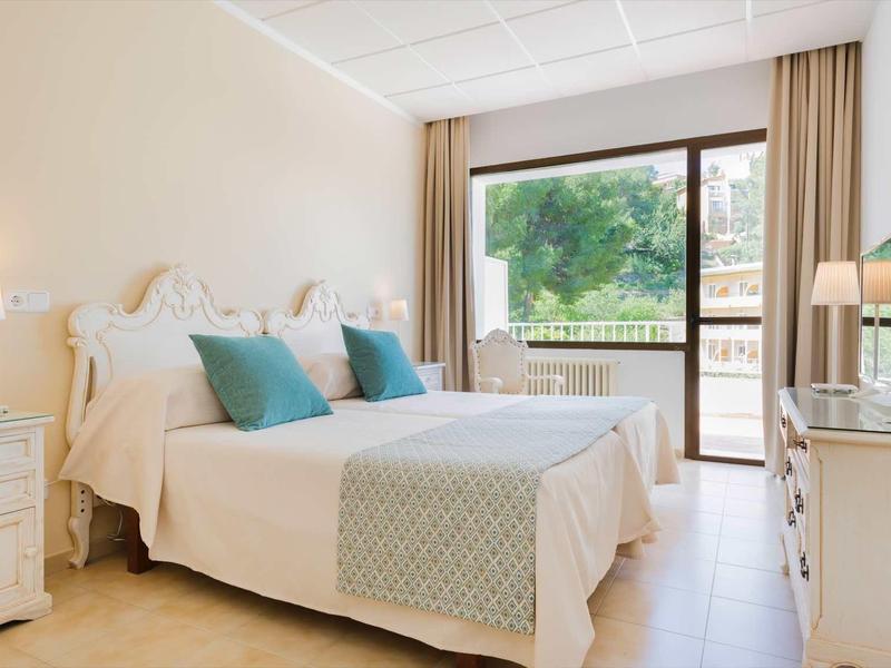 Lichte hotelkamer met tweepersoonsbed, turquoise kussens en balkon met uitzicht op groen.