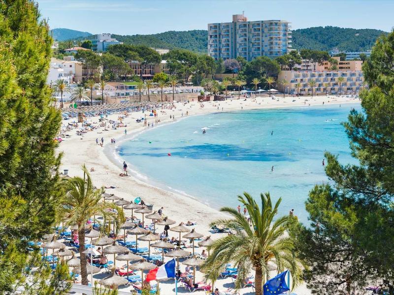 Strand met ligstoelen, palmbomen en hotels tegen beboste heuvels onder een heldere hemel.