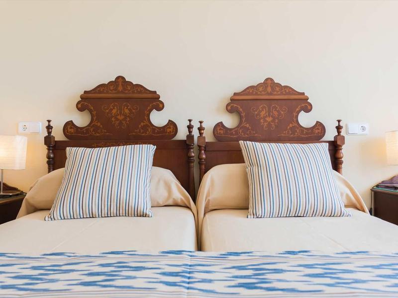 Twee eenpersoonsbedden met sierlijke houten hoofdeinden, elk met gestreepte kussens en nachtlampen.