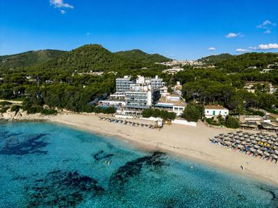 Hotel u moře s dlouhou písečnou pláží a horami v pozadí pod modrou oblohou.