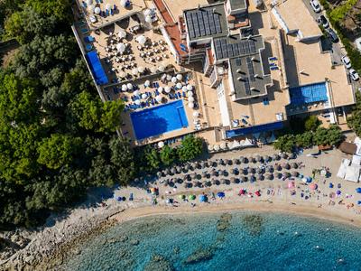 Hotel s terasou na střeše, bazénem a lehátky vedle písečné pláže a lesnaté oblasti.