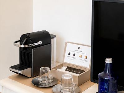 Kaffeemaschine mit zwei Tassen, Gläserverpackung und Wasserflasche auf einer Hotelzimmerablage.
