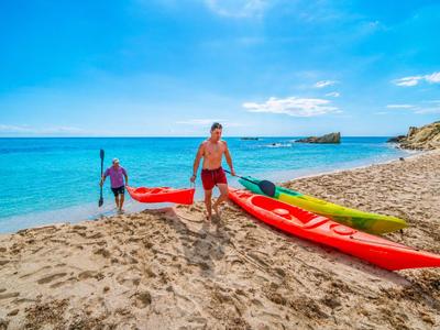 Zwei Männer bringen ein rotes Kajak an einen Sandstrand mit blauem Meer und klarem Himmel.