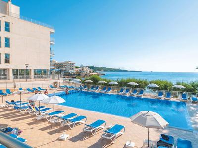 Hotelpool mit blauen Liegestühlen und Schirmen neben dem Meer unter klarem Himmel.