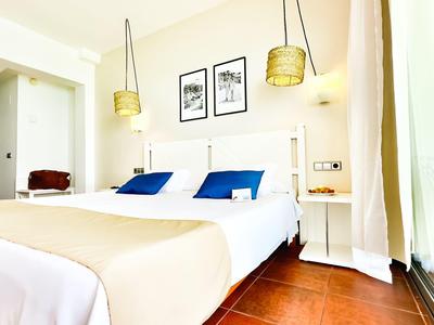 Helles Hotelzimmer mit Doppelbett, blauen Kissen und zwei Wandlampen neben einem bodentiefen Fenster.