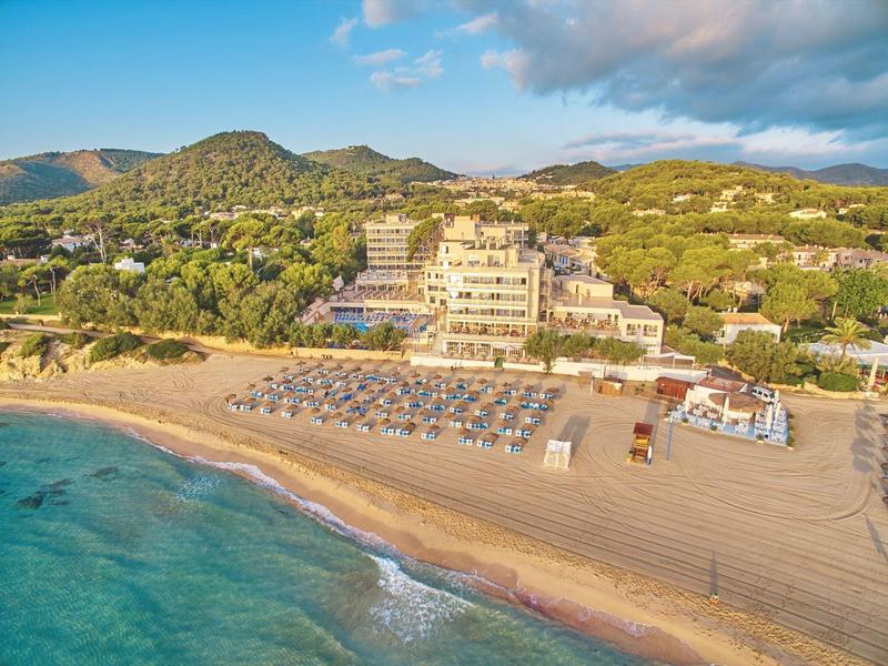 Blick auf ein großes Hotel am Strand mit Sonnenschirmen und Bergen im Hintergrund.