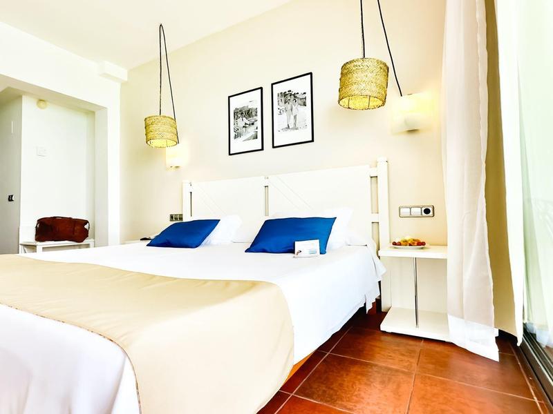 Helles Hotelzimmer mit Doppelbett, blauen Kissen und zwei Wandlampen neben einem bodentiefen Fenster.