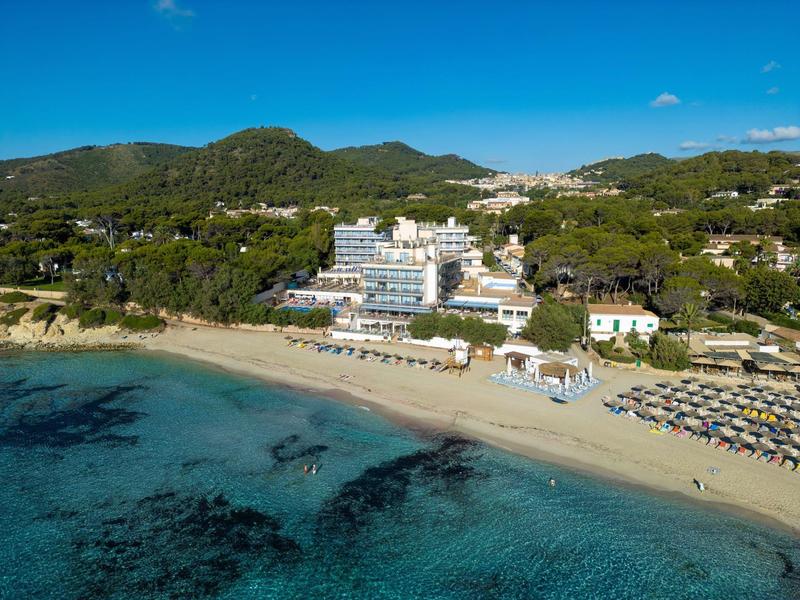 Weißes Hotel am Strand mit Bergen und klarem blauem Wasser im Hintergrund.