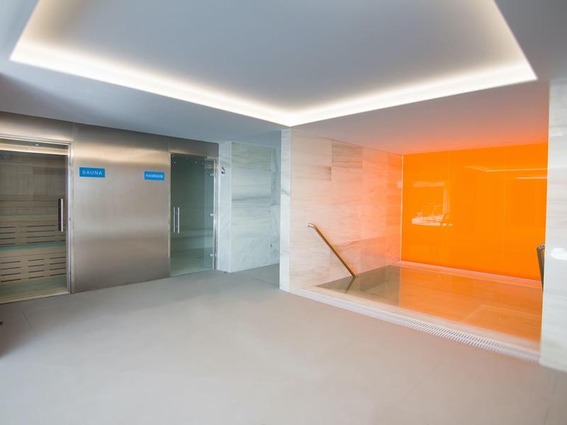 Modern hotel lobby with elevators on the left and a bright orange accent wall by stairs.