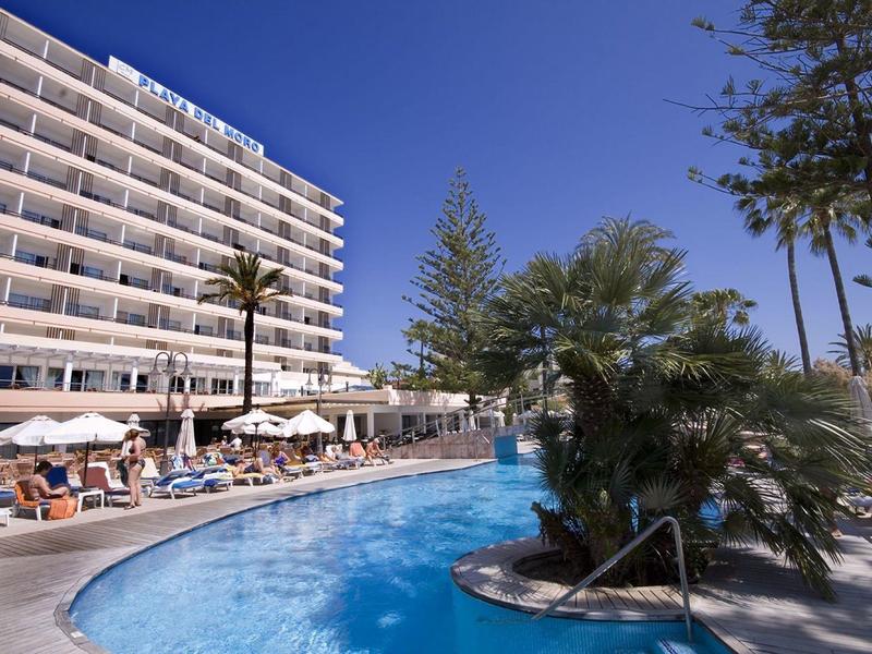 Hotel with outdoor pool, palm trees, sun loungers, and clear blue sky on a sunny day.