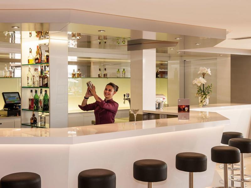 Modern hotel bar with bartender mixing a drink and stools along the bright counter.