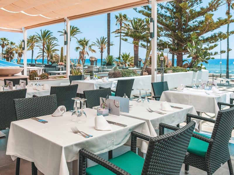 Open-air restaurant with set tables, green chairs, palm trees, and sea view.