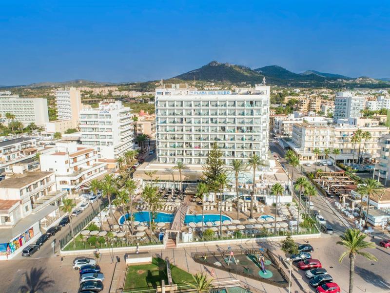 Resort hotel with pool, palm trees, and city buildings under clear blue sky.