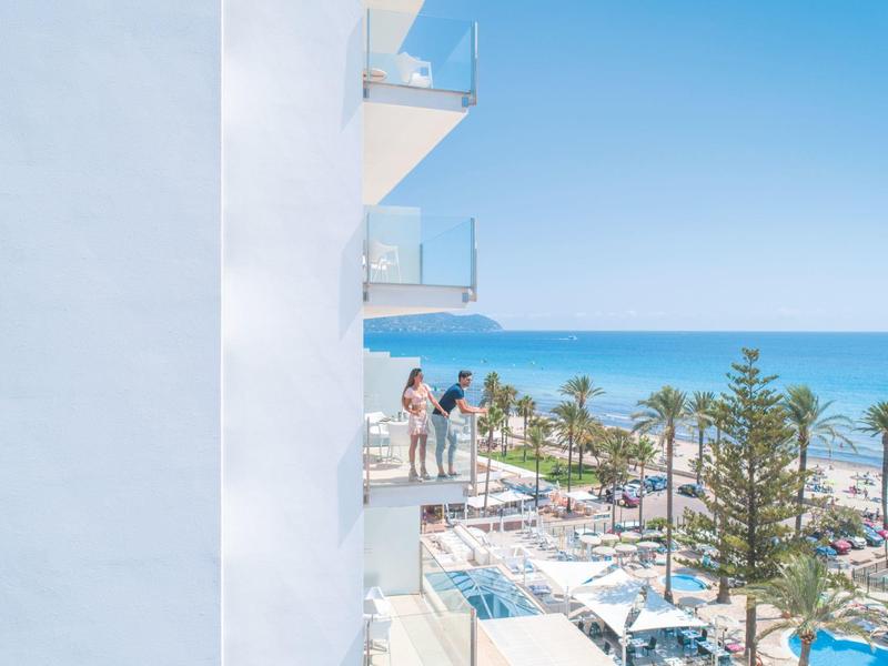 A balcony with guests on a white hotel building overlooking the sea and pool.