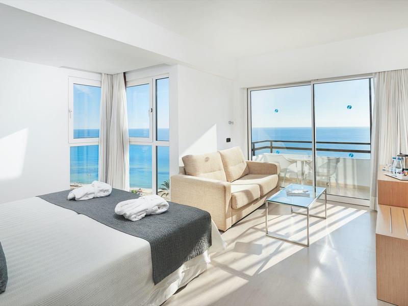 Modern hotel room with sea view, double bed, sofa, and balcony with glass railing.
