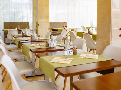 Moderner, heller Gastraum mit Holz- und weißen Möbeln, grünen Tischläufern und eingedeckten Tischen.