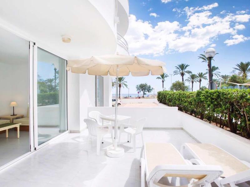 Helle Terrasse mit weißen Möbeln, Sonnenschirm und Blick auf Palmen bei blauem Himmel.