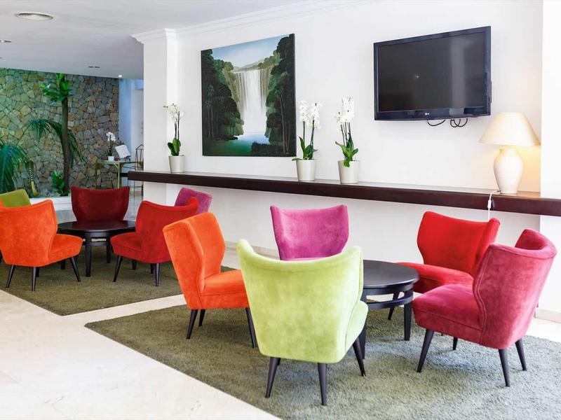 Moderner Wartebereich mit bunten, gepolsterten Sesseln, Tischen, TV und Pflanzen an weißer Wand.