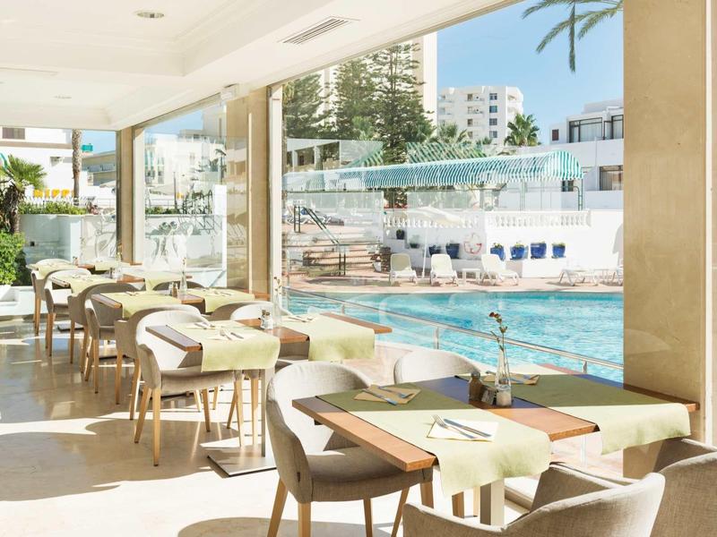 Helles Café mit Holztischen und Stühlen neben großem Fenster mit Blick auf Swimmingpool und Gebäude.