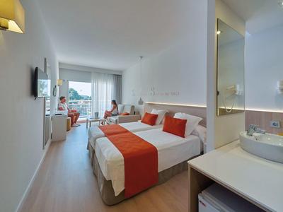 Helles Hotelzimmer mit Doppelbett, weißen Wänden, roten Akzenten und Balkon mit Meerblick.
