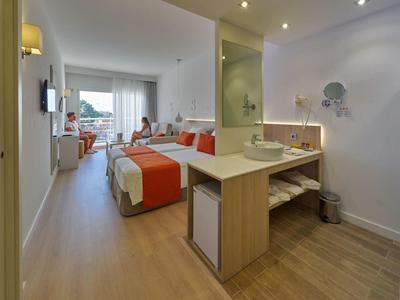 Helle Hotelzimmer mit Doppelbett, orangefarbener Bettdecke, Schreibtisch und Glas-Schiebetür zum Balkon.