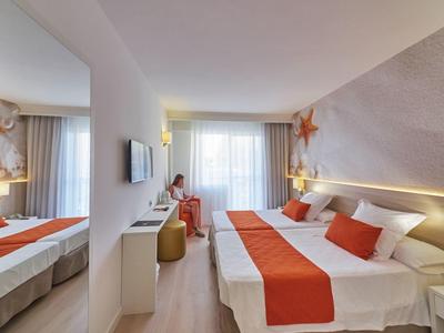 Hell eingerichtetes Hotelzimmer mit Doppelbett, orangen Akzenten und großer Fensterfront.