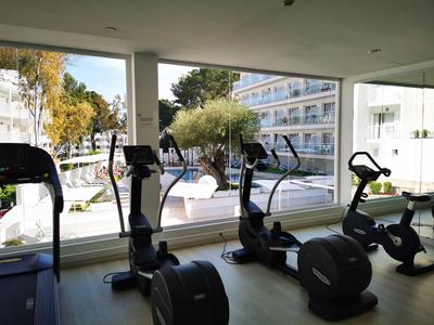 Innenraum mit drei Fitnessgeräten vor bodentiefen Fenstern, draußen Bäume und modernes Gebäude.