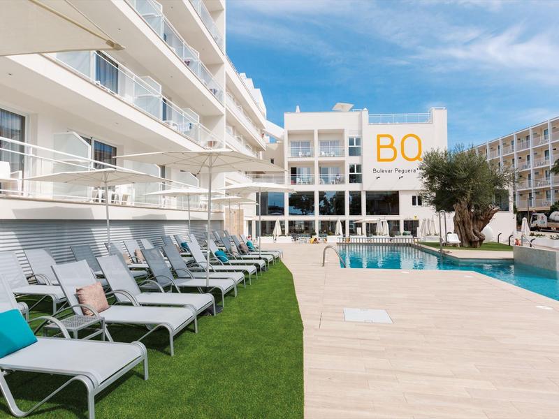 Nowoczesny hotel z basenem, leżakami i trawiastym terenem pod niebieskim niebem.