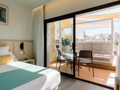 Habitación de hotel luminosa con cama doble y acceso a balcón soleado con mesa y sillas.