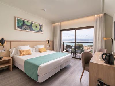 Habitación de hotel luminosa con cama doble, escritorio, balcón con vista al mar y decoración moderna.