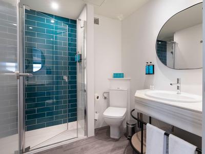 Baño de hotel moderno con ducha de vidrio, inodoro blanco, espejo redondo y lavabo sobre estante de madera.