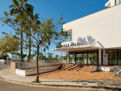Entrada moderna de hotel con terraza soleada y palmeras bajo un cielo despejado.