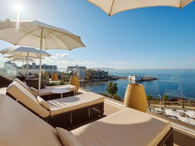 Tumbona con sombrilla en terraza con vista al mar y cielo azul despejado.