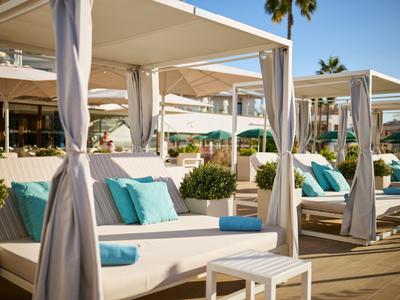 Cómodas camas lounge con cortinas blancas y cojines azules en una zona soleada de piscina del hotel.