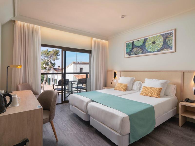 Habitación de hotel luminosa con dos camas individuales, escritorio, balcón y decoración moderna.