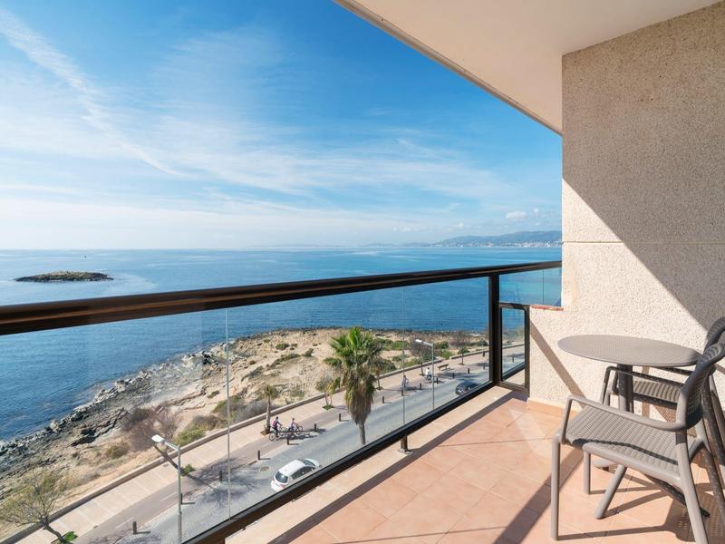 Balcón con sillas y barandilla de vidrio con vista al mar y acantilados bajo cielo azul.