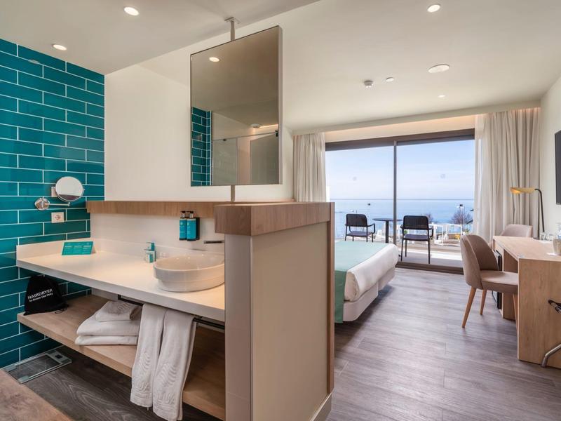 Habitación de hotel moderna con pared verde, cama doble y vista al mar desde el balcón.