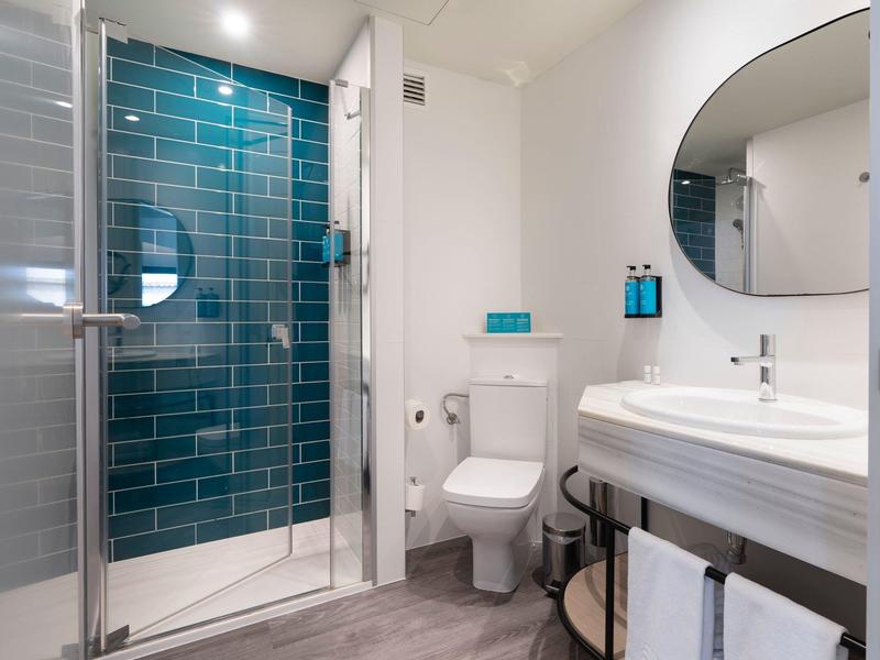 Baño de hotel moderno con ducha de vidrio, inodoro blanco, espejo redondo y lavabo sobre estante de madera.