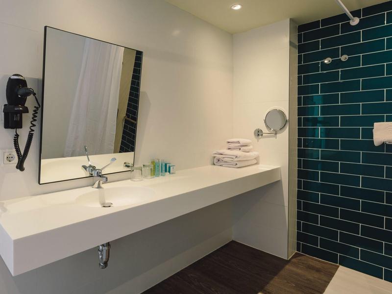 Área de baño moderna en hotel con espejo grande, lavabo, secador de pelo y azulejos azules.