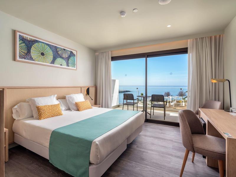 Habitación de hotel luminosa con cama doble, escritorio y vistas al mar desde el balcón.