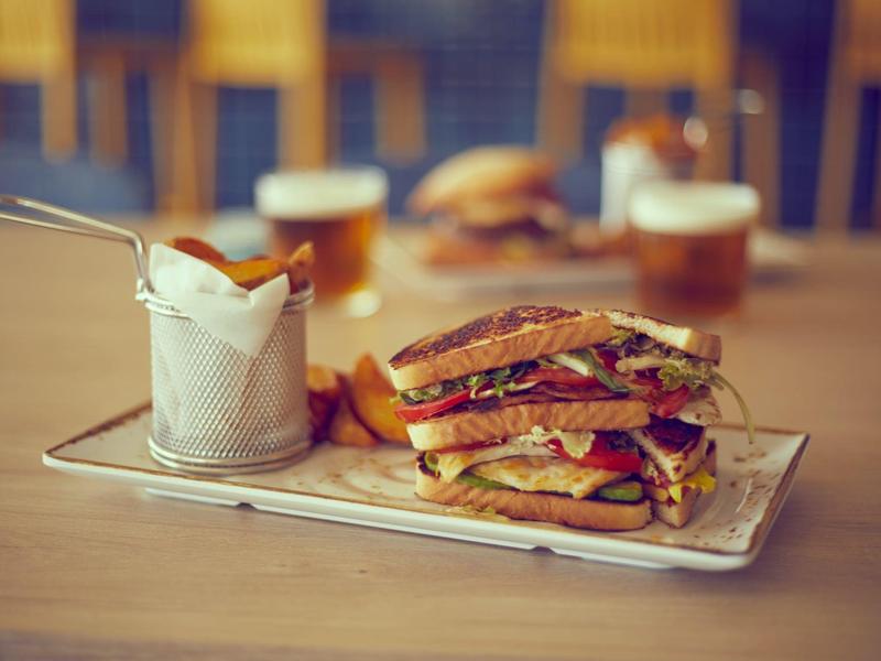 Sándwich con carne y verduras, acompañado de papas fritas y cerveza sobre una mesa de madera.