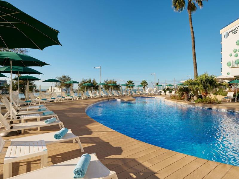 Piscina de hotel con tumbonas, sombrillas y palmeras bajo un cielo azul despejado.