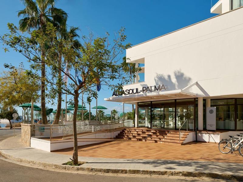 Entrada moderna de hotel con terraza soleada y palmeras bajo un cielo despejado.
