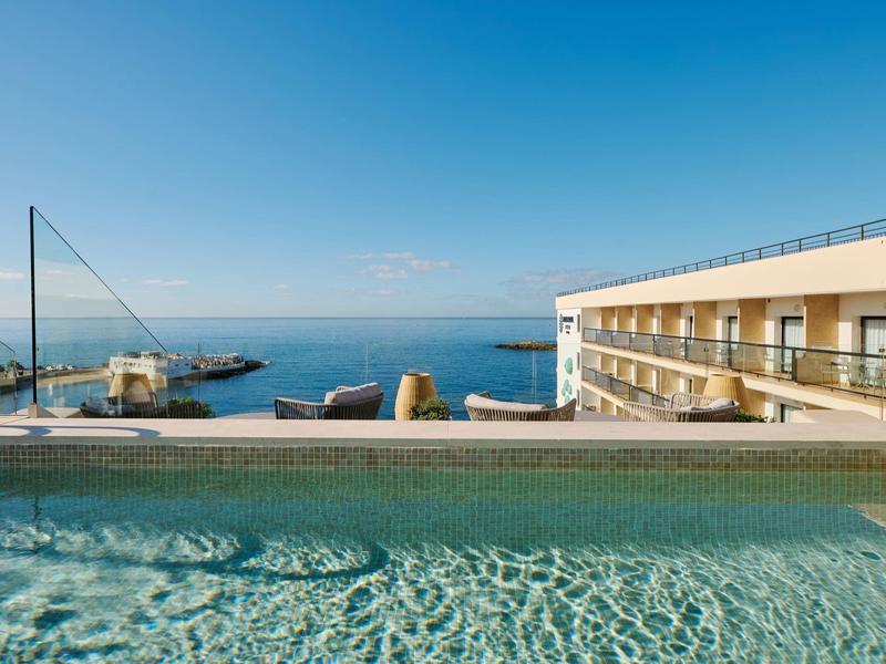 Vista de una piscina con barandilla de vidrio transparente y el mar al fondo bajo cielo despejado.