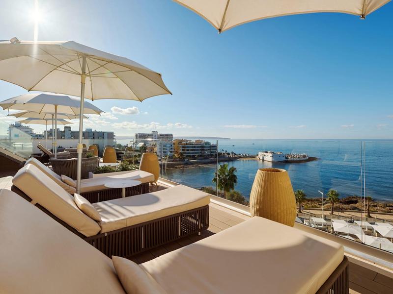 Tumbona con sombrilla en terraza con vista al mar y cielo azul despejado.