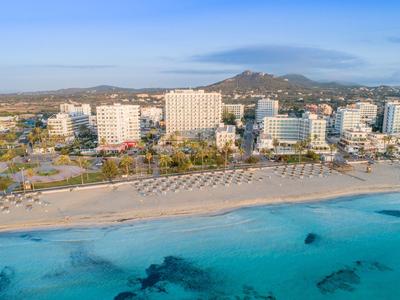 Een kustresort met hotels, een zandstrand en helderblauwe zee onder een heldere lucht.