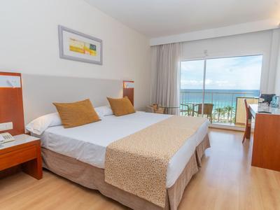 Habitación de hotel luminosa con cama doble, vista al mar a través de grandes ventanas y balcón.